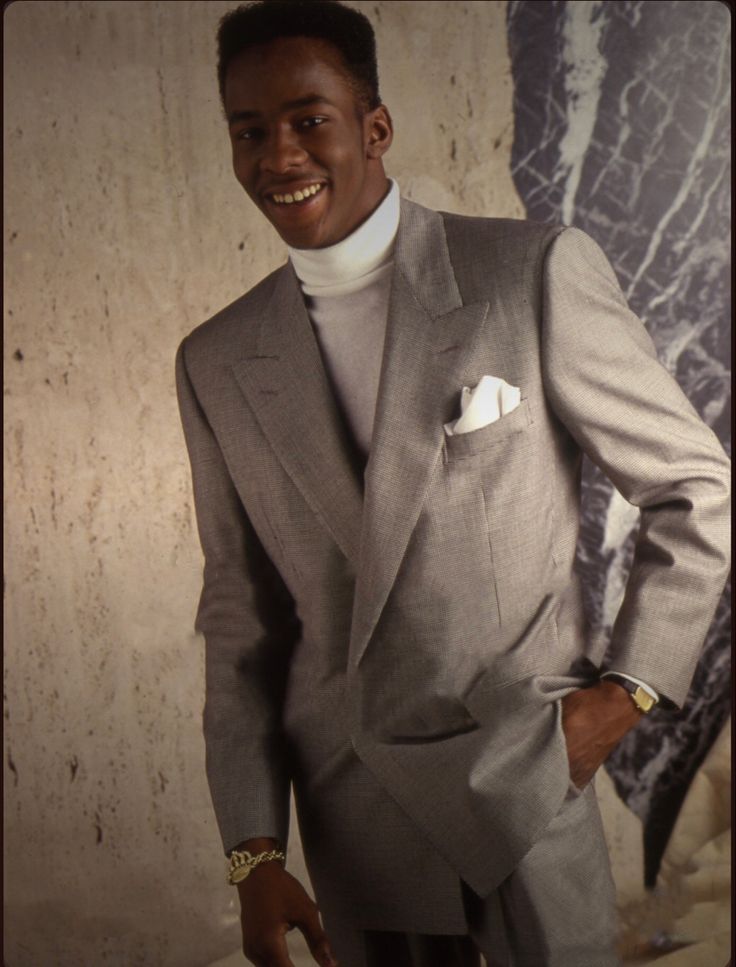 a man in a gray suit and white turtle neck sweater smiles while standing against a wall