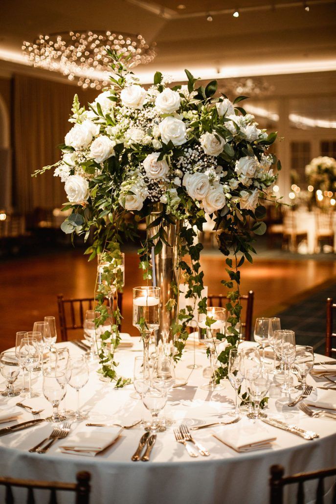 a tall vase filled with white flowers and greenery on top of a round table