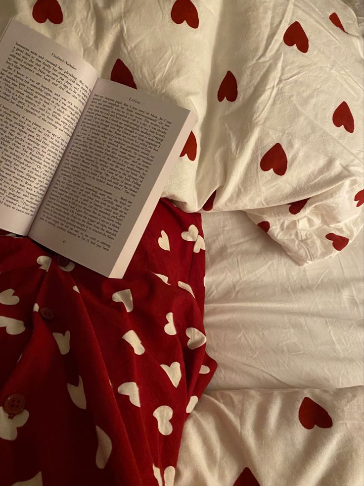 an open book laying on top of a bed next to pillows and sheets with hearts