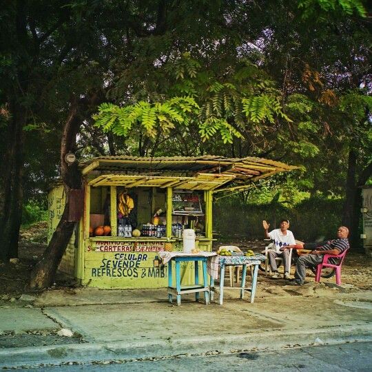 three people sitting at a table in front of a small food stand on the side of a road