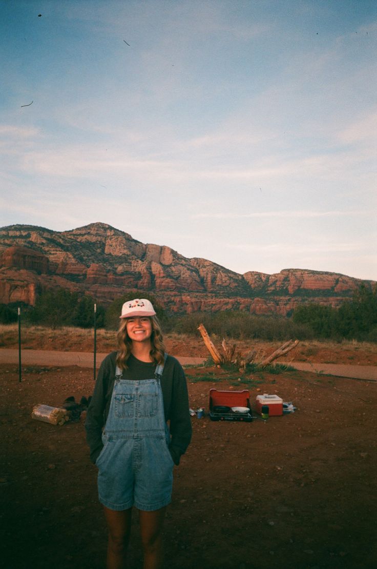 a woman wearing overalls and a white hat standing in front of mountains with tools