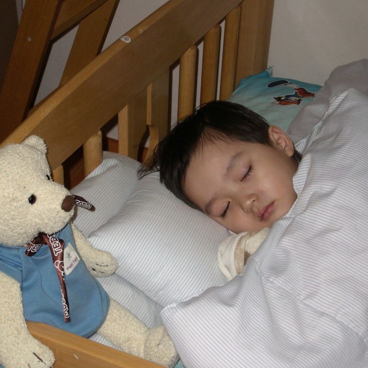 a small child sleeping in a bed next to a teddy bear and pillow with his eyes closed