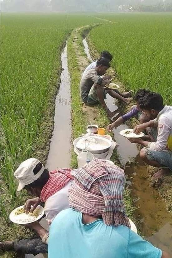 some people are sitting in the mud and eating food from bowls on their knees while another person is kneeling down behind them