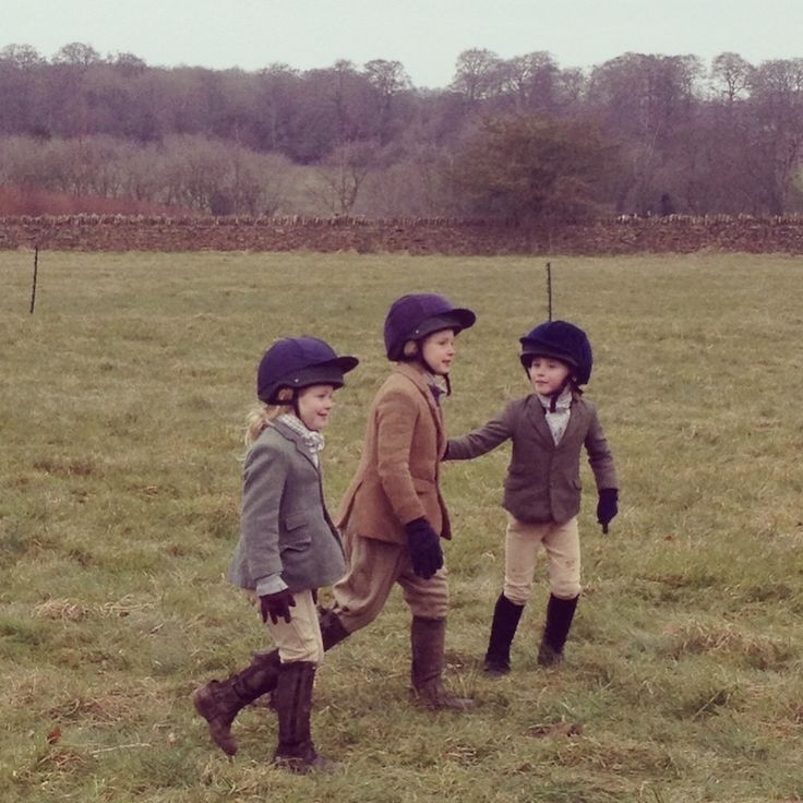 three young children in riding gear walking through a field with one holding the hand of another child's hand