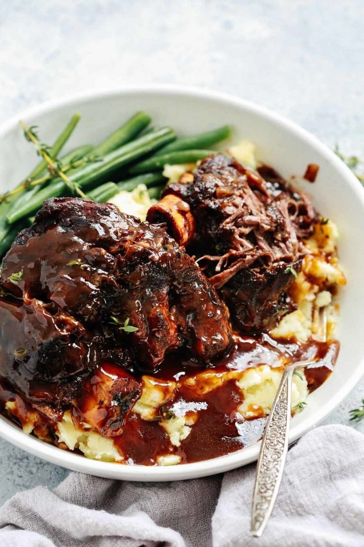 a white bowl filled with meat and potatoes covered in gravy next to green beans