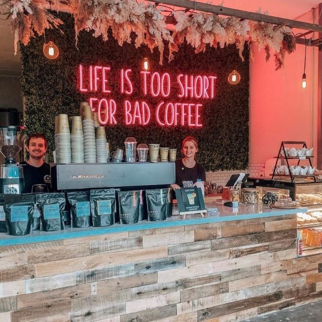 two people standing behind a counter with coffee bags on it and a neon sign that says life is too short for bad coffee