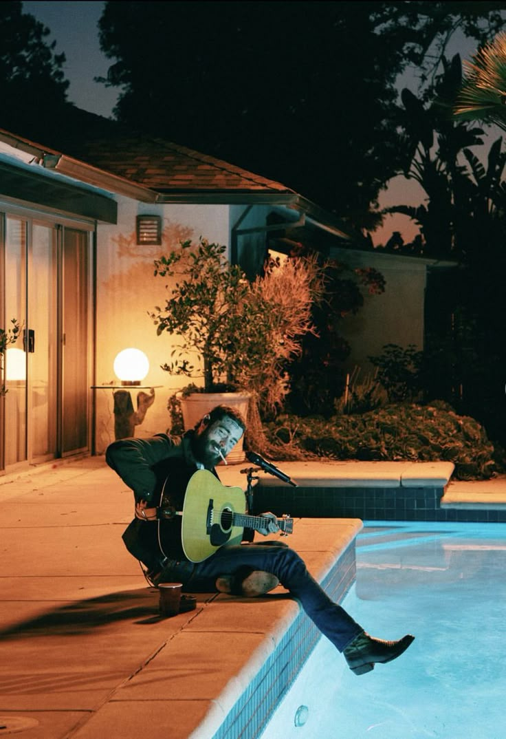 a man sitting next to a swimming pool holding a guitar