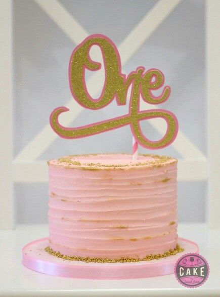 a pink and gold cake with the word one on it's top is shown