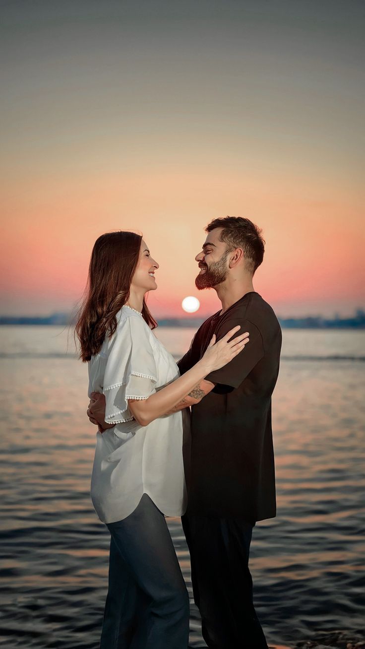 a man and woman standing next to each other in front of the ocean at sunset