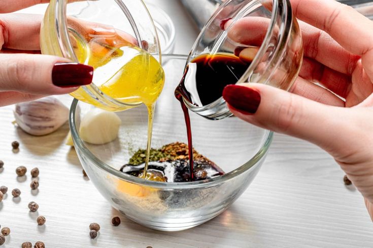 two hands pouring oil into a glass bowl filled with spices and other ingredients on a table