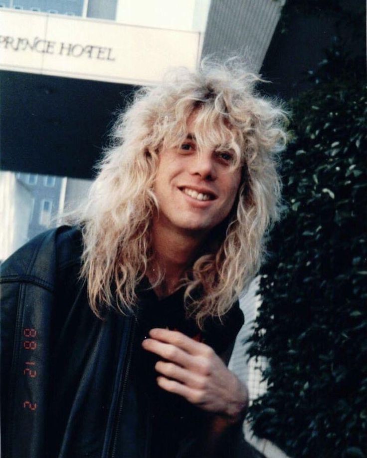 a man with long blonde hair standing in front of a building and smiling at the camera