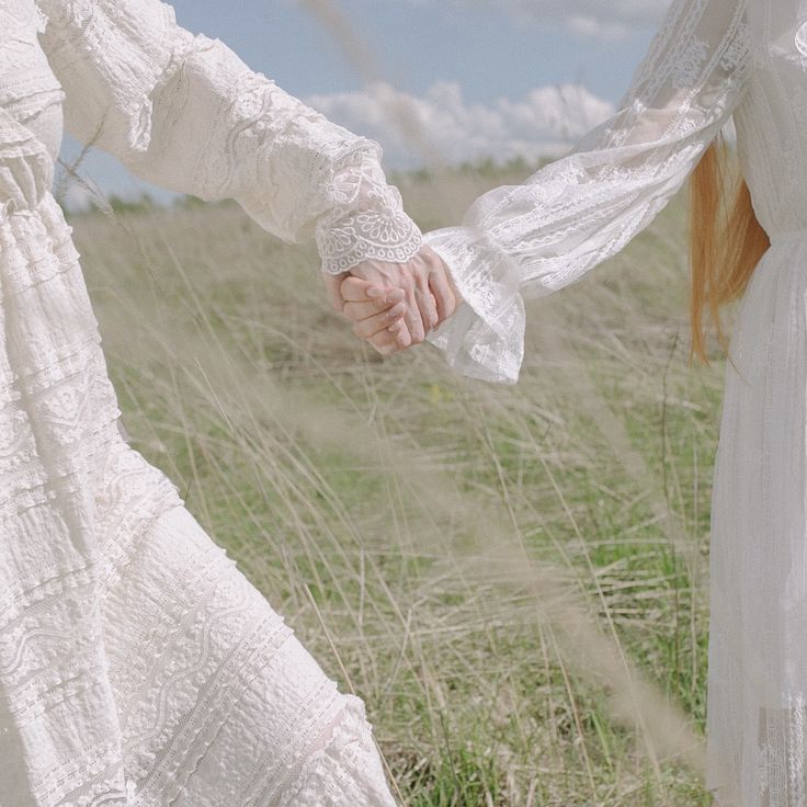 two women in white dresses holding hands and walking through tall grass on a sunny day