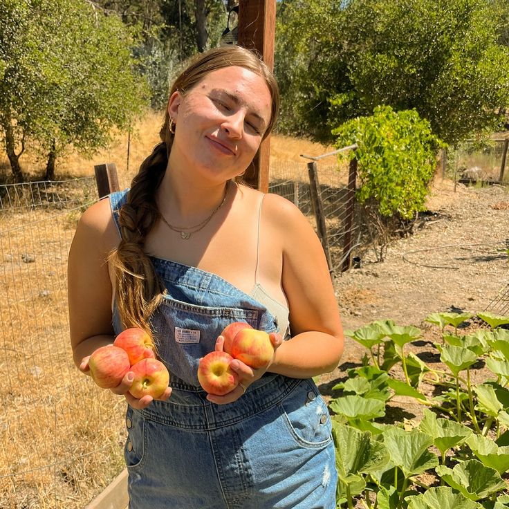 a woman in overalls holding three peaches and looking at the camera with her eyes closed