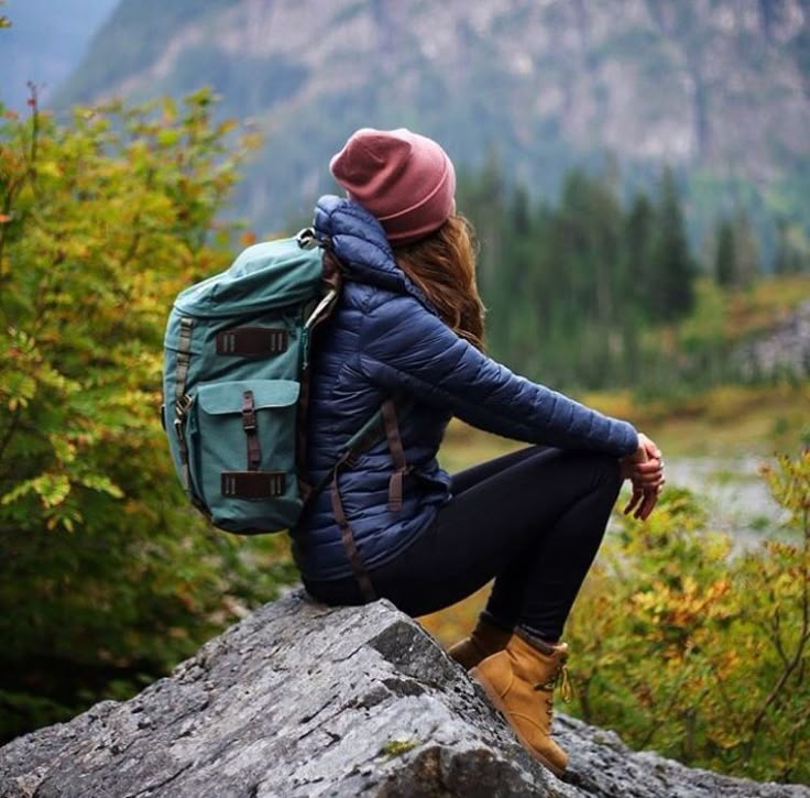 a woman sitting on top of a tree stump with a backpack over her shoulder and looking at the mountains