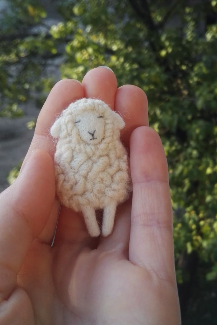 a hand holding a small white sheep in it's left palm, with trees in the background