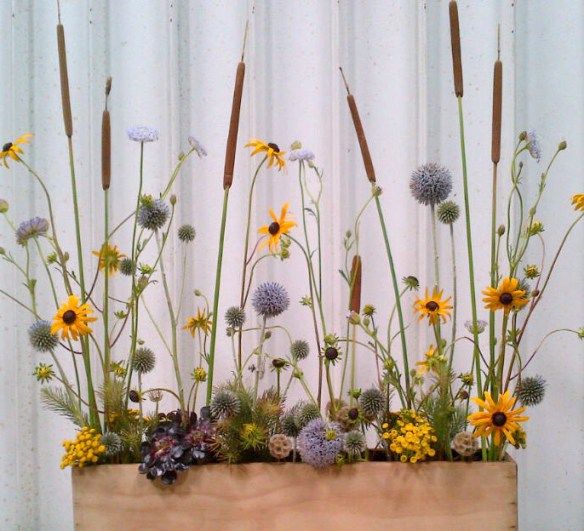 a wooden planter filled with lots of wildflowers and grasses in front of a curtain