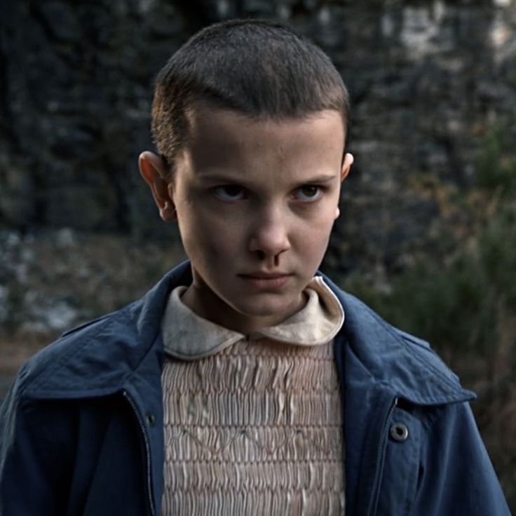 a young boy wearing a blue jacket standing in front of some bushes and trees with his head turned to the side