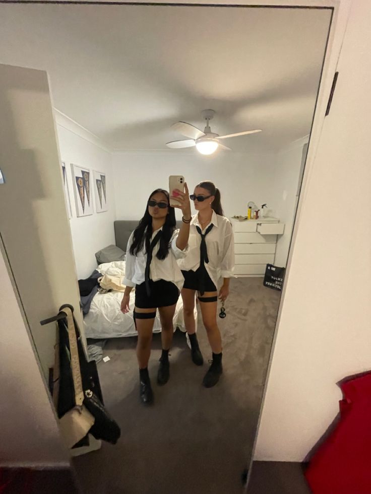 two women in short shorts are taking a selfie with their cell phone while standing in front of a mirror