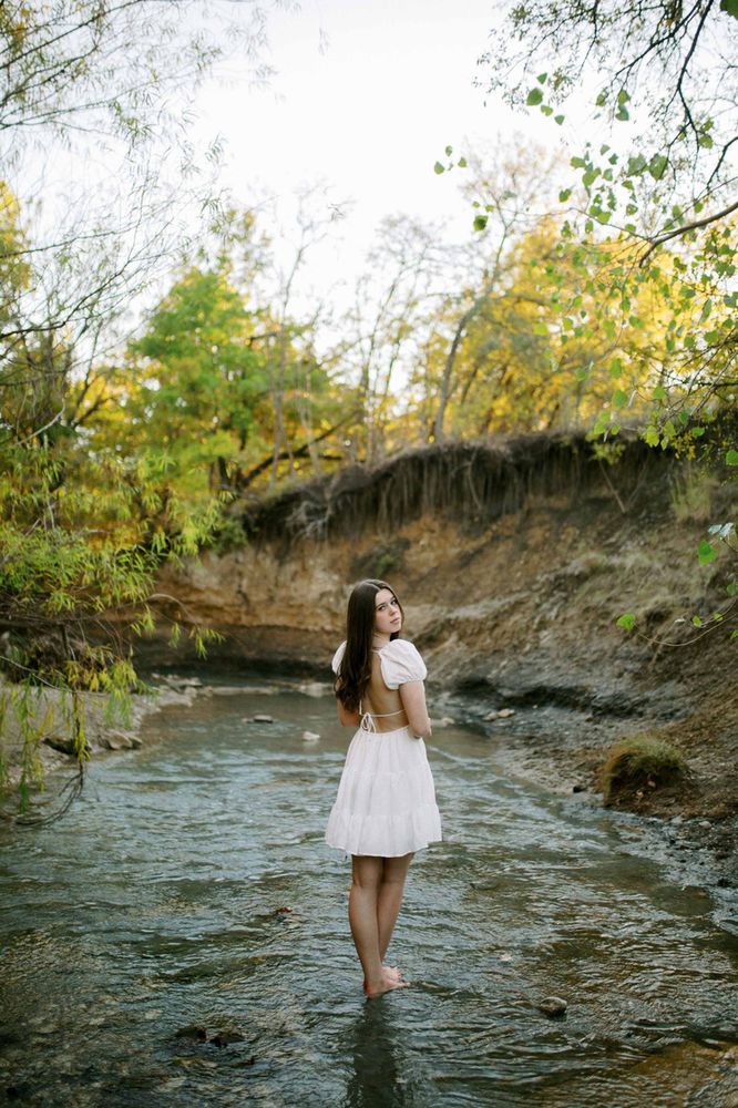 a woman in a white dress standing on the edge of a river looking up at something