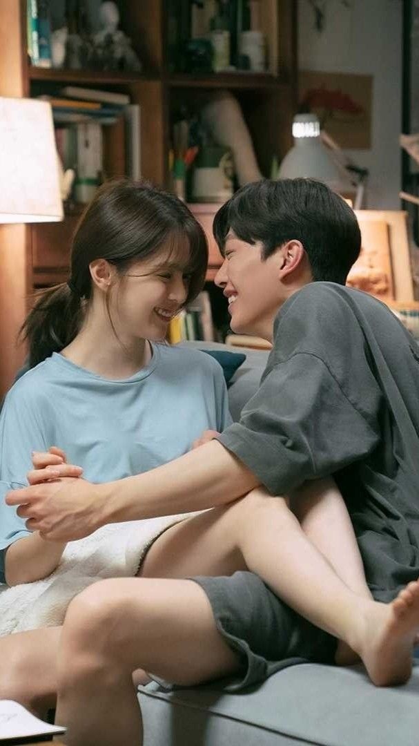 a young man and woman sitting on top of a bed next to each other in front of a bookshelf