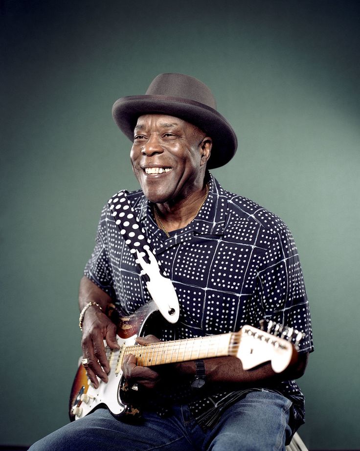 a man with a hat is playing an electric guitar and smiling at the camera while sitting on a chair