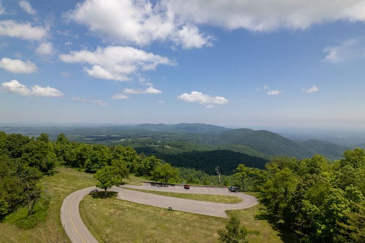 an aerial view of a winding road surrounded by trees and mountains in the distance with blue skies