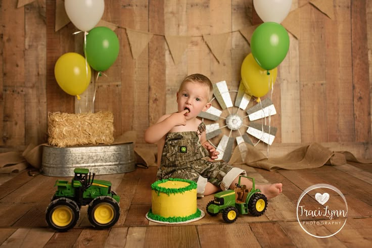 a baby boy sitting on the floor in front of a cake with tractor decorations and balloons