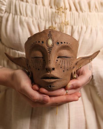 a woman is holding a clay mask in her hands, with eyes painted on it