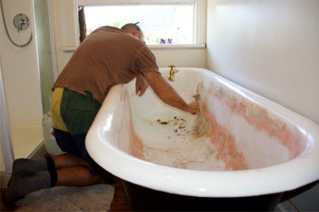 a man in brown shirt and green shorts cleaning a bathtub with red dirt on it