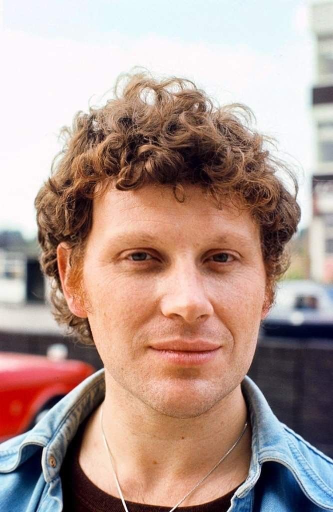 a man with curly hair wearing a blue jacket and necklace in front of a red car