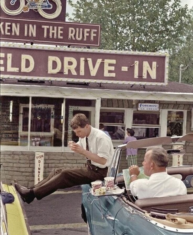 two men sitting in an old car at the drive - in, talking on their cell phones