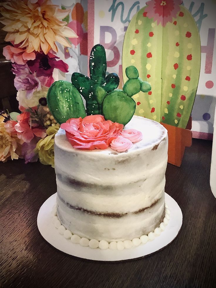 a white cake with pink flowers and cactus decorations on top, sitting on a wooden table