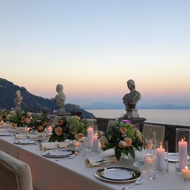 an outdoor dining table with candles and flowers on it, overlooking the ocean at dusk