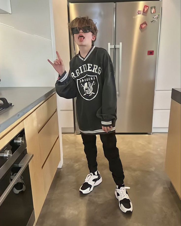 a young boy standing in the middle of a kitchen with his hand up and wearing sunglasses