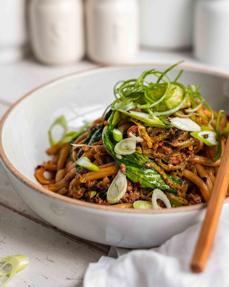 a white bowl filled with noodles and vegetables next to two wooden chopsticks on top of a table