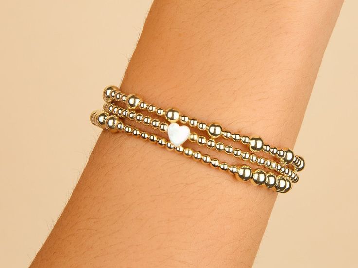 Dainty and enchanting, this 3mm bead bracelet with a Mother of Pearl heart charm is a symphony of subtle elegance. The golden beads offer a warm glow, while the heart charm brings a meaningful whisper of love and tenderness to the wrist. Ideal for layering or wearing solo, it's a versatile token of affection that's as timeless as it is trendy.﻿ 14K gold filled. Bead size: 3mm. Mother of Pearl Heart Charm: 6.5 x 7mm. 6.75 inch strong stretchy elastic band. Hypoallergenic, waterproof and tarnish r Bead Heart, Golden Beads, Pearl Heart, Subtle Elegance, Heart Bracelet, Heart Charm Bracelet, A Mother, Bead Bracelet, Gold Beads