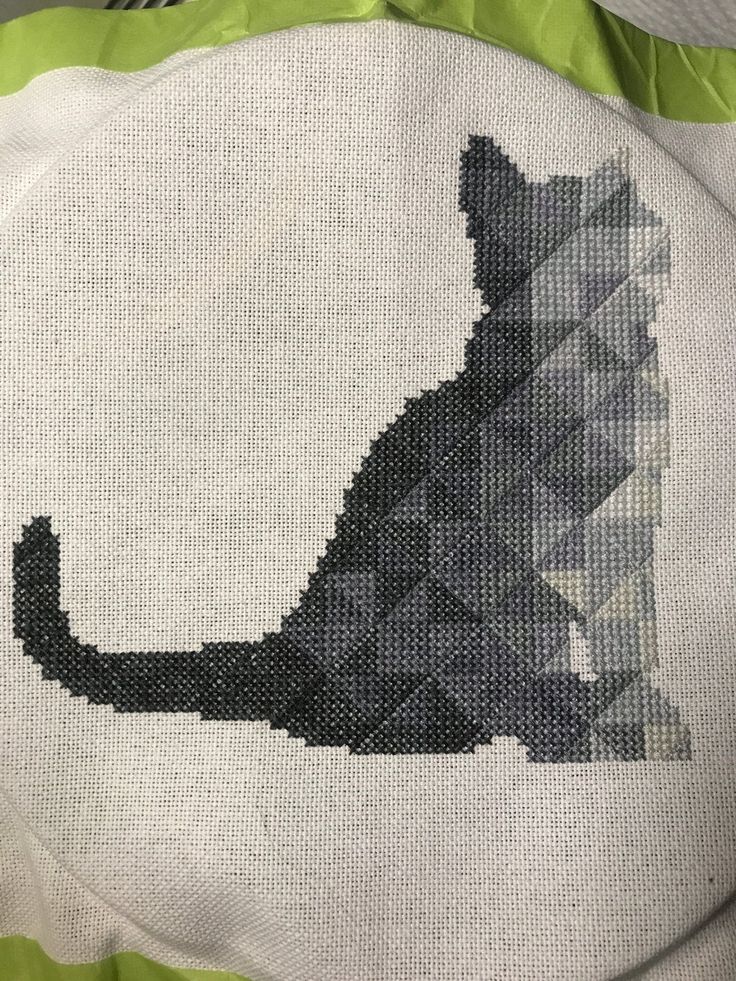 a cross stitch cat sitting on top of a green and white pillow with the silhouette of a cat