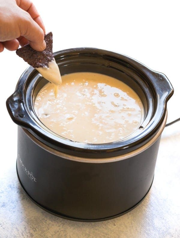 a hand holding a cracker over a pot of white queso dip