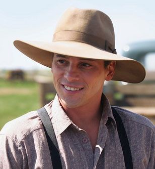 a man wearing a hat and suspenders smiles at the camera while standing in front of a fence