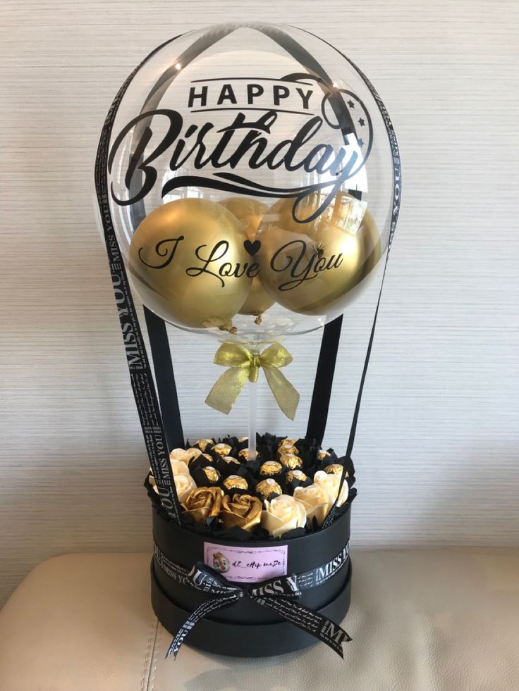 a birthday cake with gold balloons and chocolates in a glass dome on a table