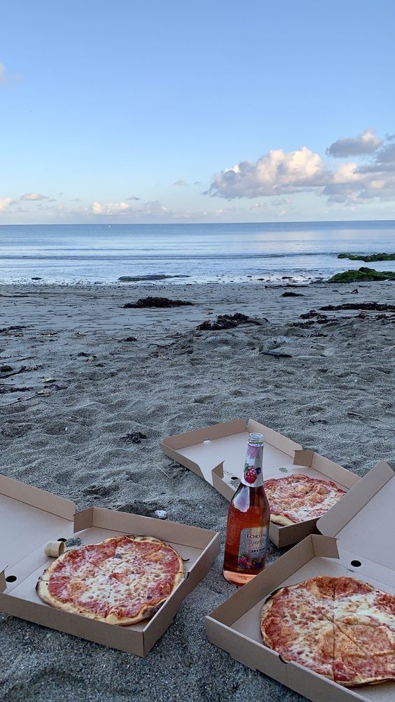 three pizzas in boxes on the beach with a bottle of beer