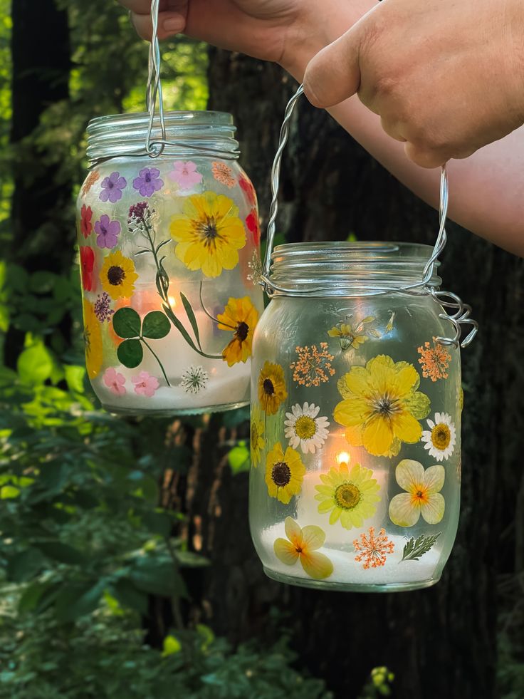 two mason jars with flowers painted on them hanging from strings in front of some trees