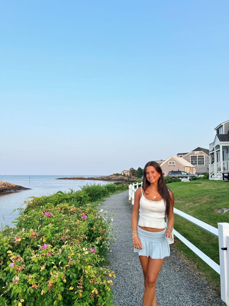 a beautiful young woman standing on top of a gravel road next to the ocean with houses in the background