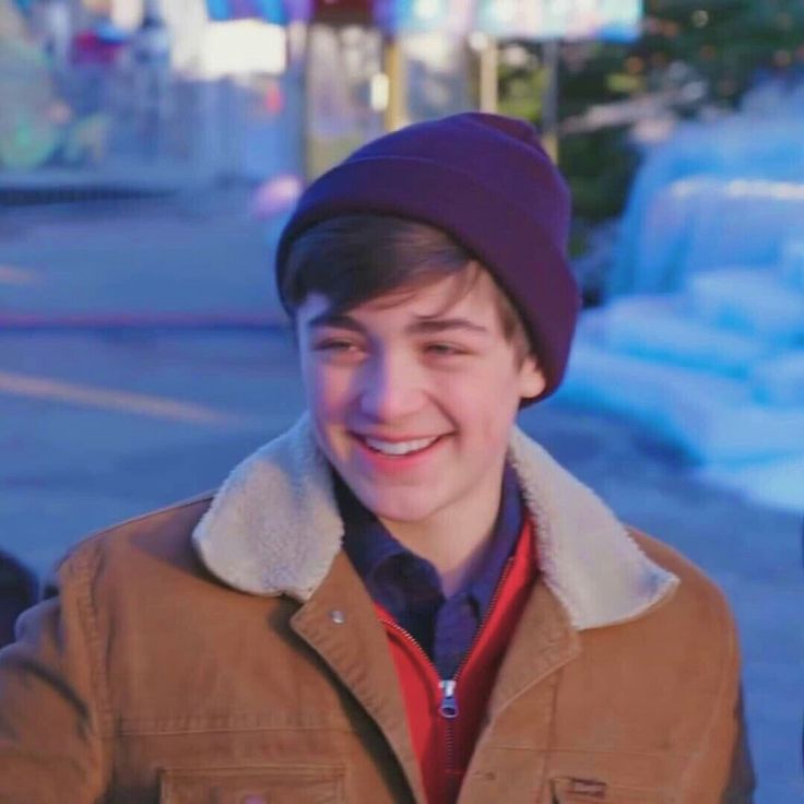 a young man wearing a brown jacket and a purple hat smiles at the camera while standing on a sidewalk