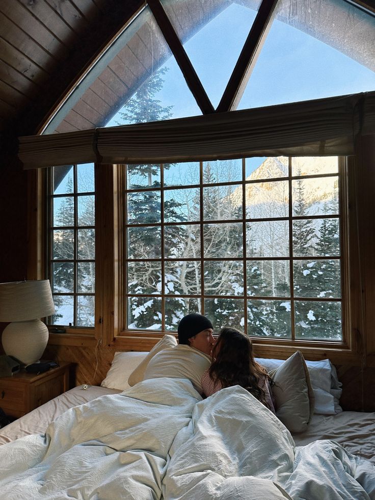 a man and woman laying in bed under a window with snow on the mountain outside