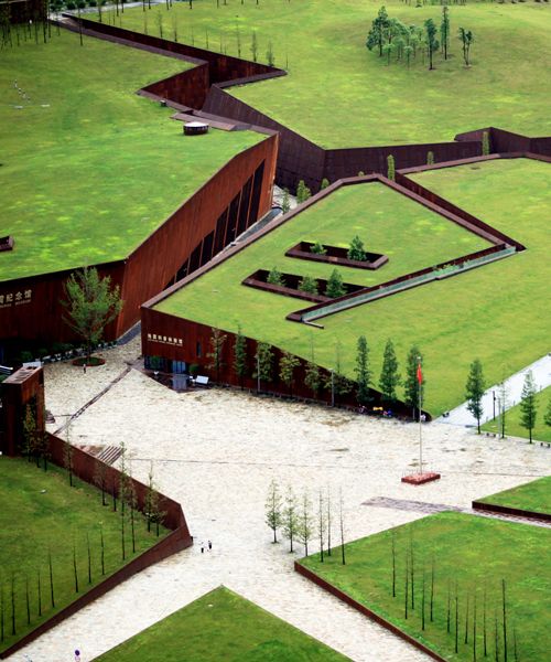 an aerial view of a green field with buildings and cars parked on the side of it