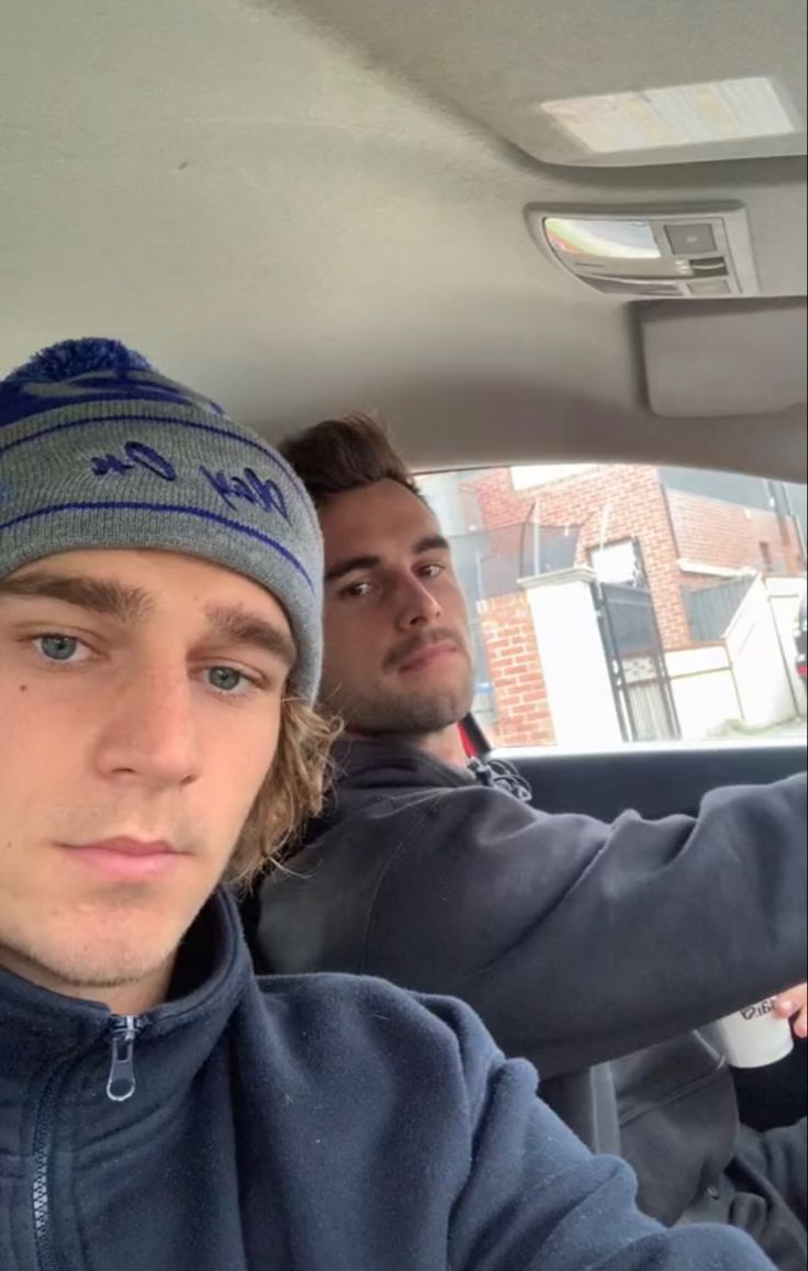 two men sitting in the back seat of a car, one wearing a beanie