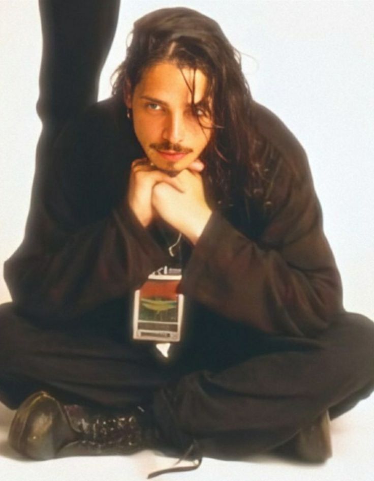 a man sitting on the ground with his hands under his chin, wearing black pants and a brown jacket