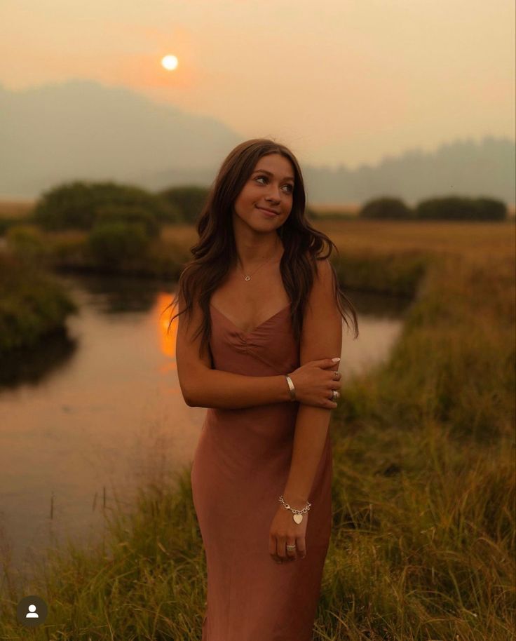 a woman in a long dress is standing by the water at sunset with her arms crossed