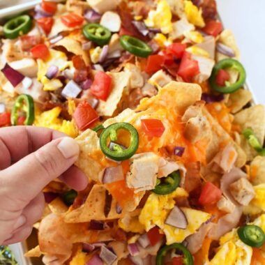 a person is reaching for a nacho with cheese and peppers on it in front of a tray of tortilla chips